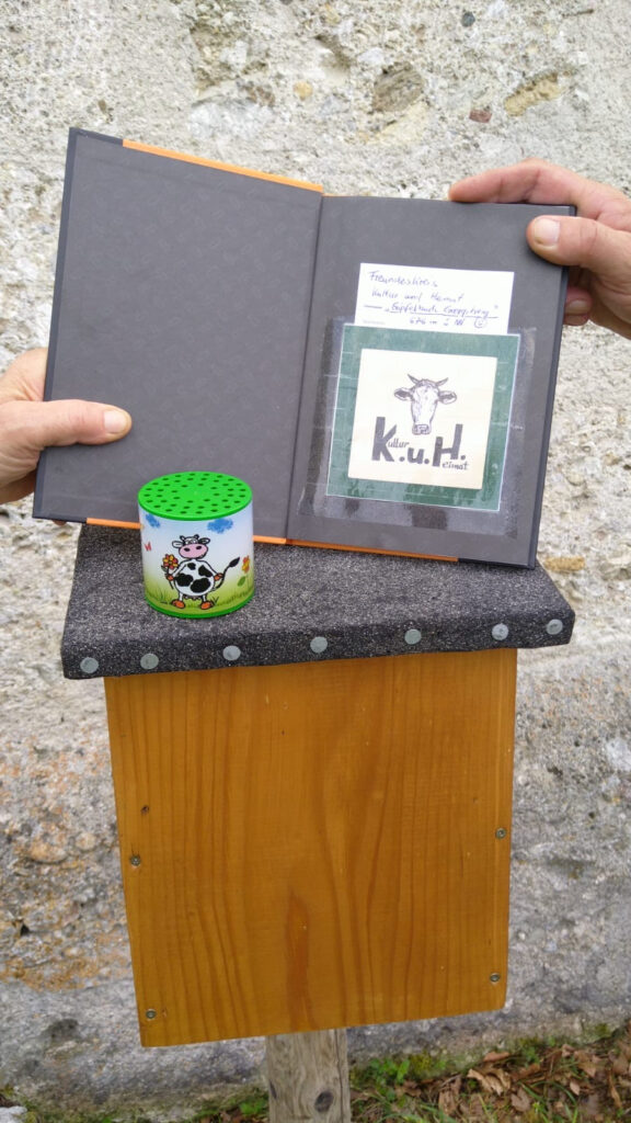Das geöffnete Gipfelbuch mit K.u.H.-Zeichen über seinem Holzkasten an der Kirchenmauer.