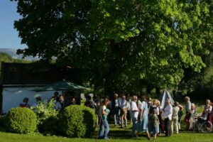 Besucher der Maiandacht stehen unter der großen Linde in Thalham; im Hintergrund sind Berge zu sehen.