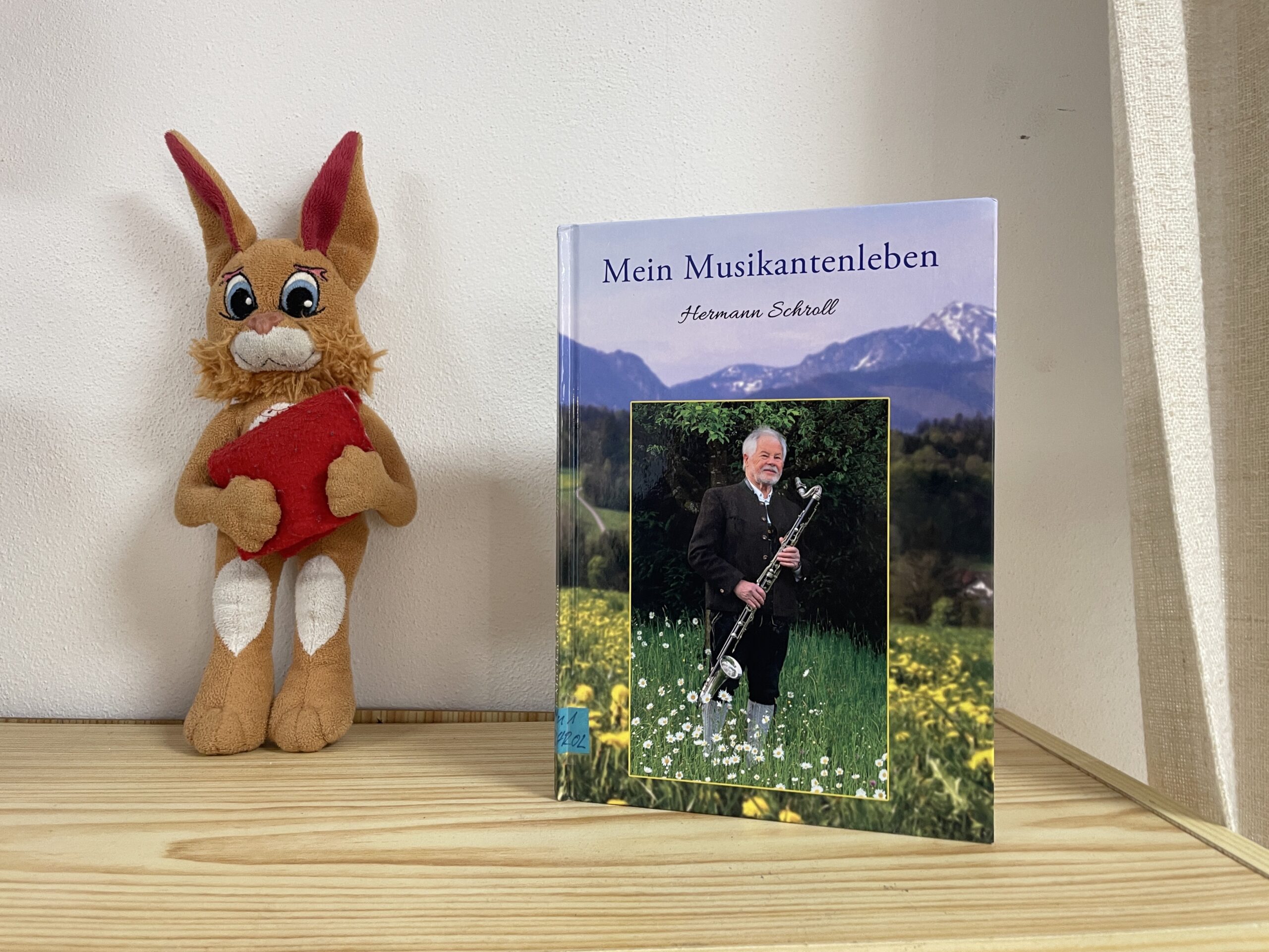 Das Buch Mein Musikantenleben von Hermann Schroll neben einem Bücherei-Maskottchen.