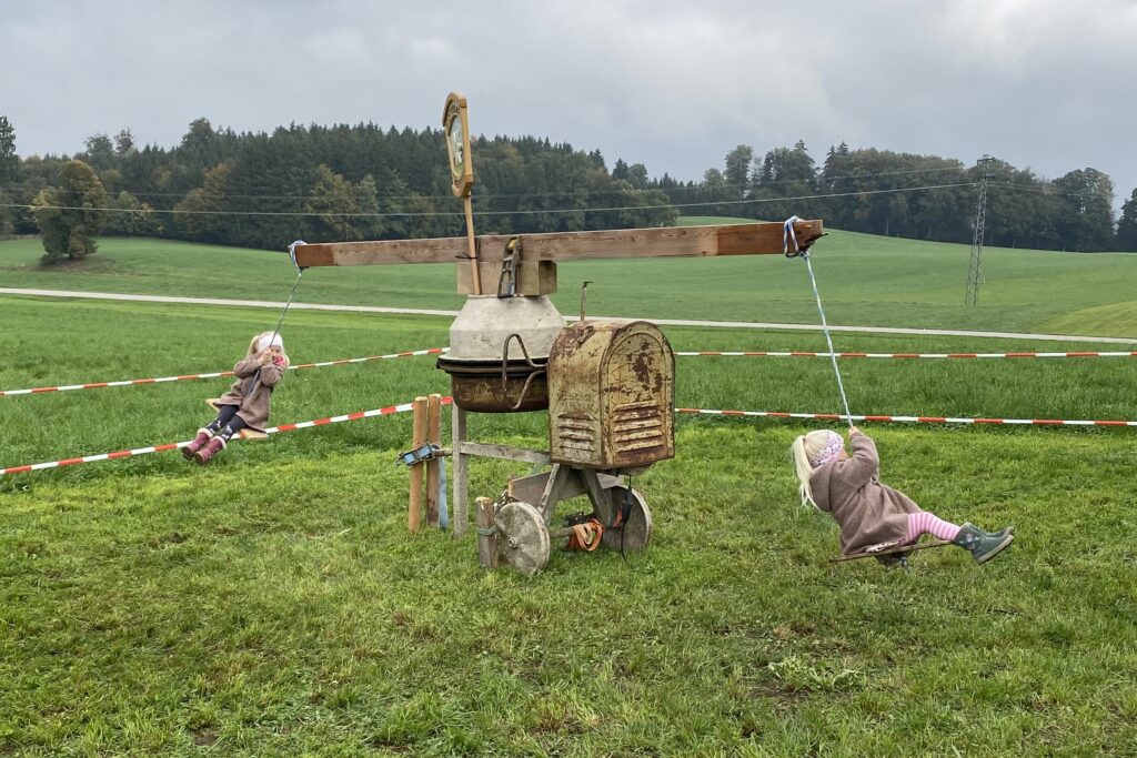 Zwei Kinder schwingen auf den Seilsitzen eines Karussells mit hölzernem Querbalken.