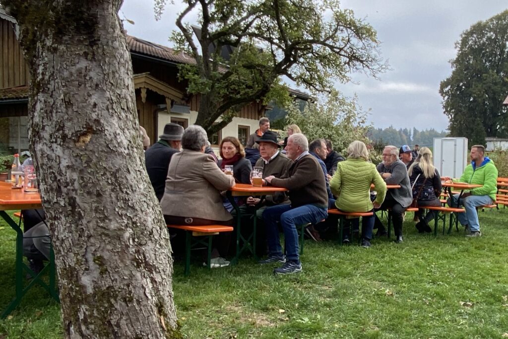 Gäste sitzen an Biertischen unter Bäumen beim Hof.