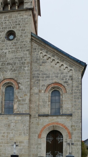 Die Frontansicht der Grabenstätter Pfarrkirche St. Maximilian zeigt mit Rundbogenportal, roten Ziegelrahmungen und Zahnschnittfries einen klaren neoromanischen Stil, der Kraft und Einfachheit ausstrahlt. 1836 von Gottfried von Neureuther entworfen, war sie damals ein "ultramodernes" Gebäude. Neuromanische Bauweise Grabenstaett st maximilian ausseneindruck