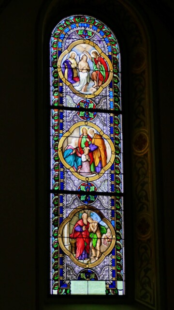 Das figürliche Fenster erzählt in drei Medaillons Szenen aus dem Leben Jesu und Mariens, eingebettet in ein leuchtend ornamentiertes Rahmenwerk. Grabenstaett st maximilian fenster