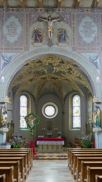 Der Chorraum beeindruckt durch seine reiche Ausmalung mit goldbetonten Ornamenten, flankierende Heiligenfiguren. Über dem Triumphbogen eine Kreuzigungsdarstellung. Grabenstaett st maximilian gesamteindruck