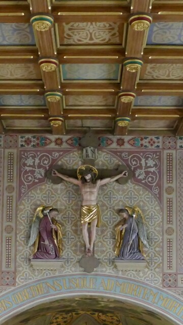 Die bemalte Kassettendecke von St. Maximilian zeigt stilisierte florale und geometrische Motive in sanften Tönen, die mit goldenen Zierleisten gefasst sind und dem Raum rhythmische Struktur verleihen.
Die Fresken von Max Fürst darunter entfalten in reichem Ornament und fein nuancierter Farbgebung ein harmonisches Zusammenspiel aus Liturgiesymbolik und dekorativer Pracht. Grabenstaett st maximilian gesamteindruck