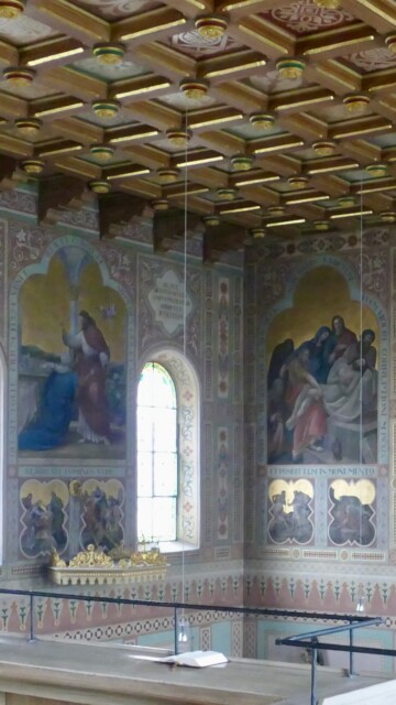 Die Kassettendecke wird von kräftigen Balken getragen und gliedert sich in Felder mit vergoldeten Rosetten und stilisierten Ornamenten – ein Muster strenger Harmonie.
Darunter die Wandfresken in leuchtender Farbigkeit und klarer Komposition, meisterlich eingebettet in reiche Rahmenornamente, die Schriftbänder und Bibelszenen malerisch verbinden. Grabenstaett st maximilian gesamteindruck