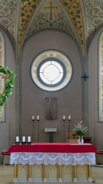 Der Hauptaltar von St. Maximilian ist schlicht gestaltet, mit goldverzierten Säulchen und rotem Altartuch, vor einer Apsis, die von feinem Rankenwerk und einem Kreuzfresko über dem Rundfenster geprägt ist. Grabenstaett st maximilian hauptaltar