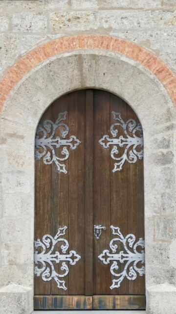 Das Außenportal von St. Maximilian besticht durch seinen Rundbogen mit Ziegelrahmung und die reich verzierten schmiedeeisernen Beschläge auf der dunklen Holztür. Grabenstaett st maximilian portal aussen