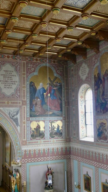 Die Fresken in St. Maximilian sind eingebettet in ein streng gegliedertes Wandornament, das die biblischen Szenen – etwa Christus mit dem Kreuz – in reicher Farbigkeit und Goldakzentuierung hervorhebt, während die Kassettendecke darüber mit goldenen Rosetten und vegetabilen Mustern eine feierliche Deckenkulisse bildet. Grabenstaett st maximilian wandbild