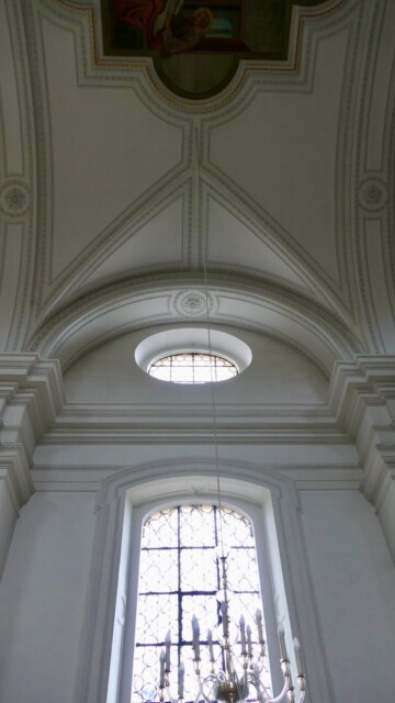 Ein hohes Rundbogenfenster durchbricht die strenge Gliederung der Wand und wird von einem ovalen Oberlicht (Occulus oder Ochsenauge) sowie fein gegliederten Stuckbändern überfangen – ein klarer, lichter Ausdruck des späten Barocks. Stichkappe mit Occulus und Fenster